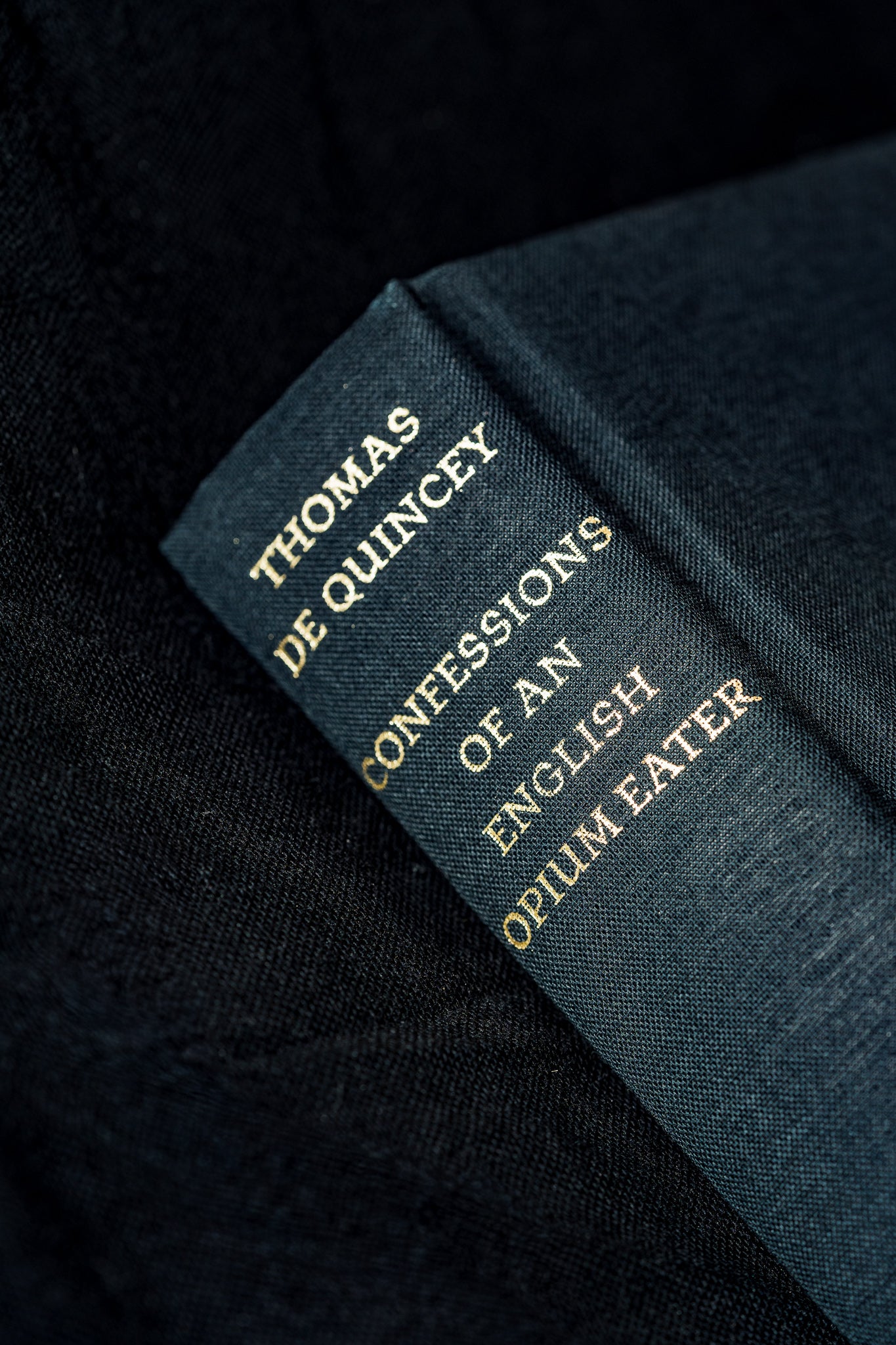 A close-up of a dark blue book spine with gold lettering reading 'THOMAS DE QUINCEY CONFESSIONS OF AN ENGLISH OPIUM EATER' on a dark textured fabric background.