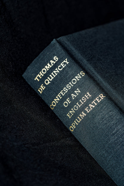 A close-up of a dark blue book spine with gold lettering reading 'THOMAS DE QUINCEY CONFESSIONS OF AN ENGLISH OPIUM EATER' on a dark textured fabric background.